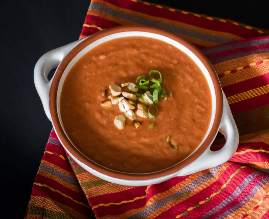 a bowl of peanut tomato soup with peanuts and chopped green onions on a red and orange napkin