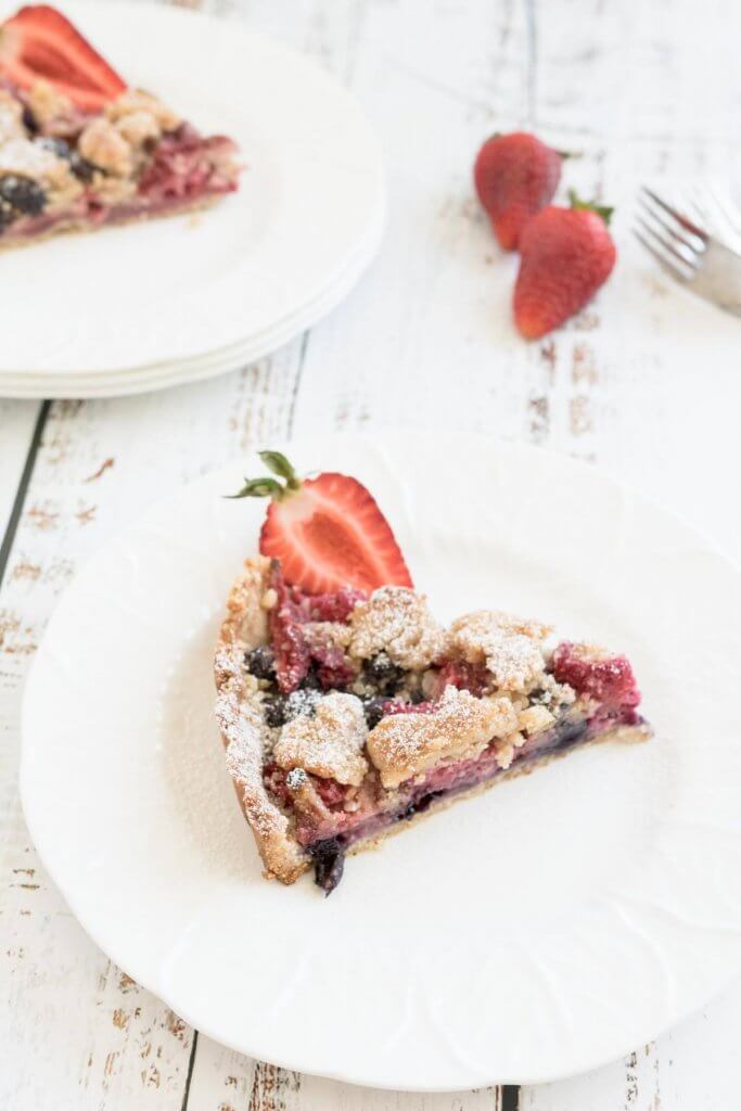 a slice of berry crumble pie on a white plate with fresh strawberries in the background.