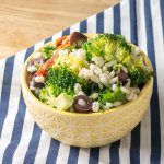 small yellow bowl on a blue striped napkin with broccoli, barley and olive salad