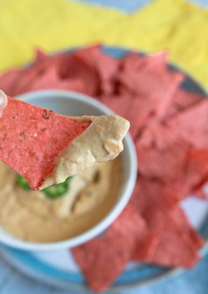 a red corn chip with vegan queso cheese dip hovering of a bowl of queso