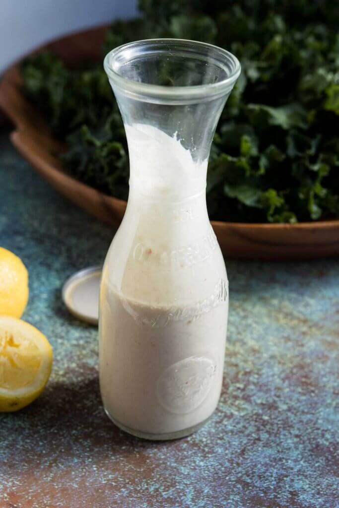 A bottle of homemade lemon tahini dressing in a glass bottle with kale salad and lemon