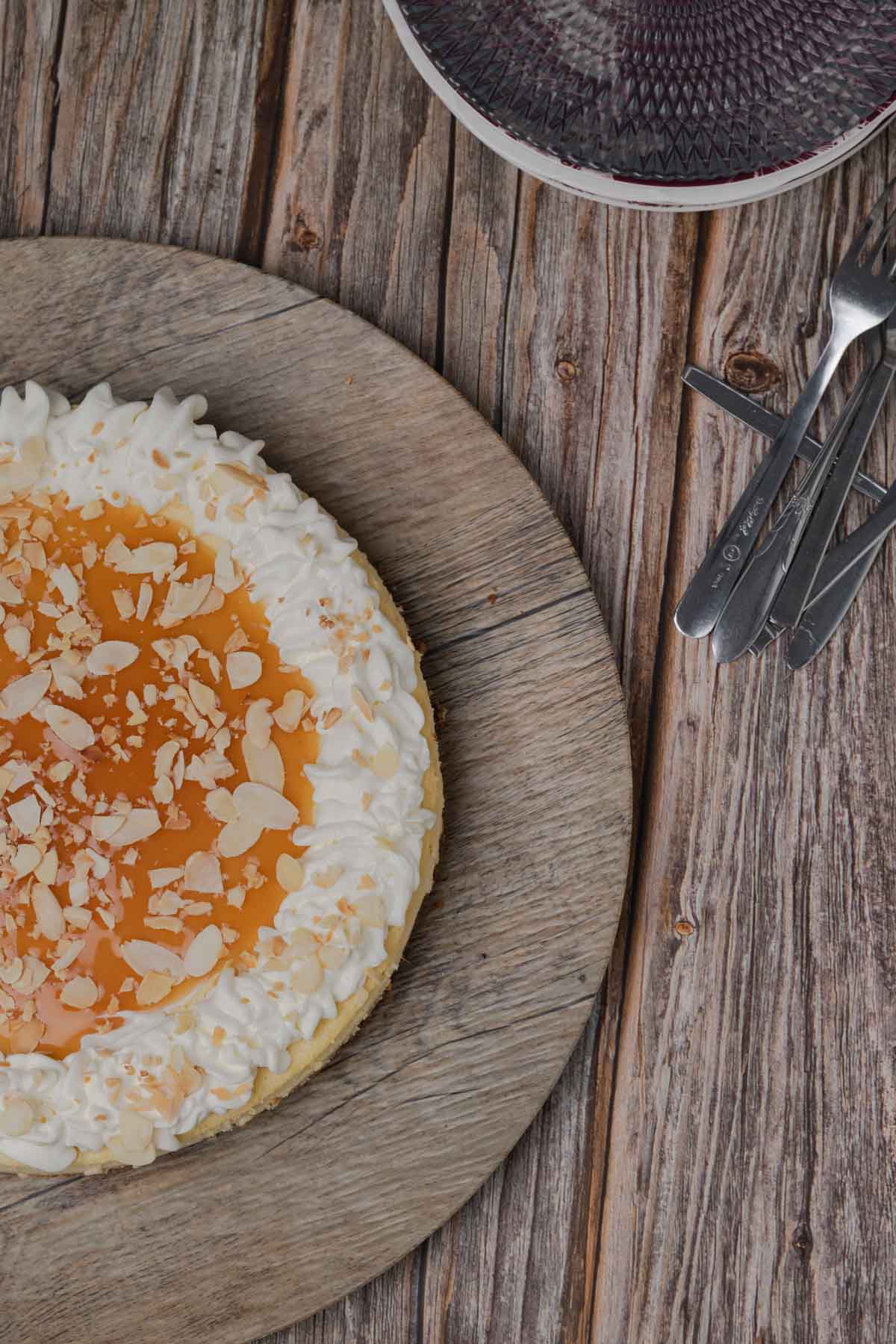 almond praline cheesecake on a wooden serving plater, cake is topped with caramel sauce, sliced almonds and almond flavoured whipped cream