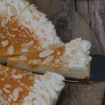 almond praline cheesecake sliced on a wooden serving plater, drizzled with caramel and almonds, one slice being served